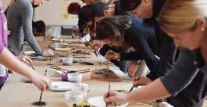 A group of people leaning over a long table, painting with brushes and ink on large sheets of paper during a collective creative workshop