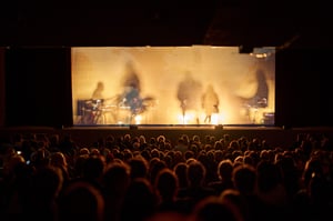 Audience seated in the theater during Telmo Pievani's show Nomadic. On stage, behind a veil illuminated by warm lights, the silhouettes of the musicians and performers can be glimpsed against the light.