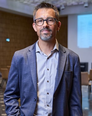 Ritratto di un relatore in giacca blu nella sala dell’evento, con lo schermo della presentazione sullo sfondo.