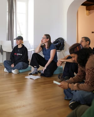 Participants sitting in a circle on cushions in a bright room, during a moment of listening and group discussion at Casa Carlo Cattaneo, Switzerland.