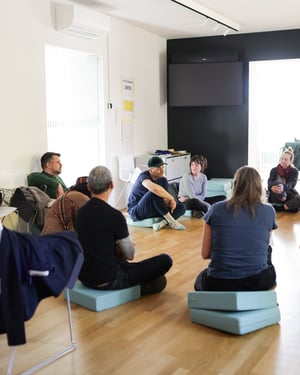 Participants sitting in a circle on cushions in a bright room, during a moment of listening and group discussion at Casa Carlo Cattaneo, Switzerland.