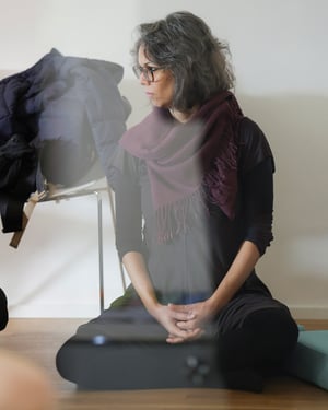 Woman sitting on a cushion, wearing a burgundy scarf and glasses, looking thoughtful during a group meeting in a bright room.