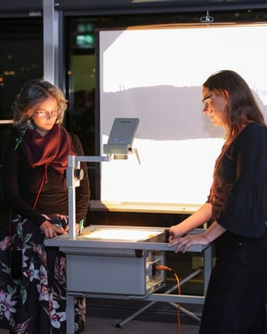 Due donne consultano un retroproiettore acceso durante una presentazione; sullo sfondo uno schermo con una proiezione luminosa in un ambiente museale serale.