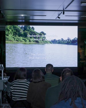 Pubblico visto di spalle mentre assiste alla proiezione di un video dedicato al fiume Tapajós; sullo schermo compaiono sottotitoli in portoghese e inglese.