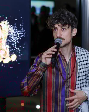 Primo piano di un relatore che parla al microfono durante un evento serale; alle sue spalle uno schermo con un’immagine luminosa.