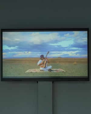 Schermo orizzontale che proietta un video di una persona seduta su un tappeto nel paesaggio desertico mentre suona una chitarra, sotto un cielo ampio e nuvoloso.