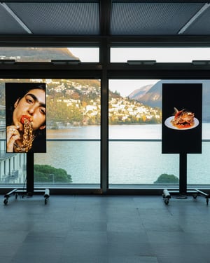 Due schermi verticali esposti davanti a una grande vetrata con vista sul lago e sulle montagne di Lugano; sulle immagini, un ritratto fotografico e una composizione artistica su fondo nero.