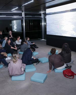 Pubblico seduto su cuscini azzurri in uno spazio museale contemporaneo mentre assiste alla proiezione di un video su un grande schermo; l’immagine mostra la superficie dell’acqua riflessa nella sala attraverso ampie vetrate.