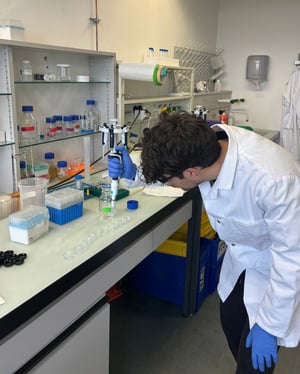 Un ricercatore con occhiali di protezione osserva l'interno di una stampante 3D con copertura arancione, in un laboratorio dotato di attrezzature e banchi di lavoro.