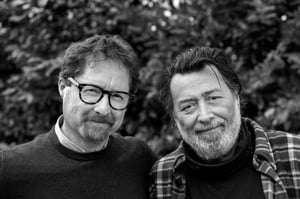 Black and white photo of two men, Telmo Pievani on the left and Gianni Maroccolo on the right, shown side by side from the waist up, both smiling.