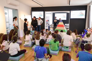 Bambini seduti su cuscini azzurri in una sala moderna e luminosa, partecipano a un laboratorio educativo con adulti che guidano la lezione. Sullo sfondo, due schermi mostrano una slide con la scritta “Torniamo alla piramide